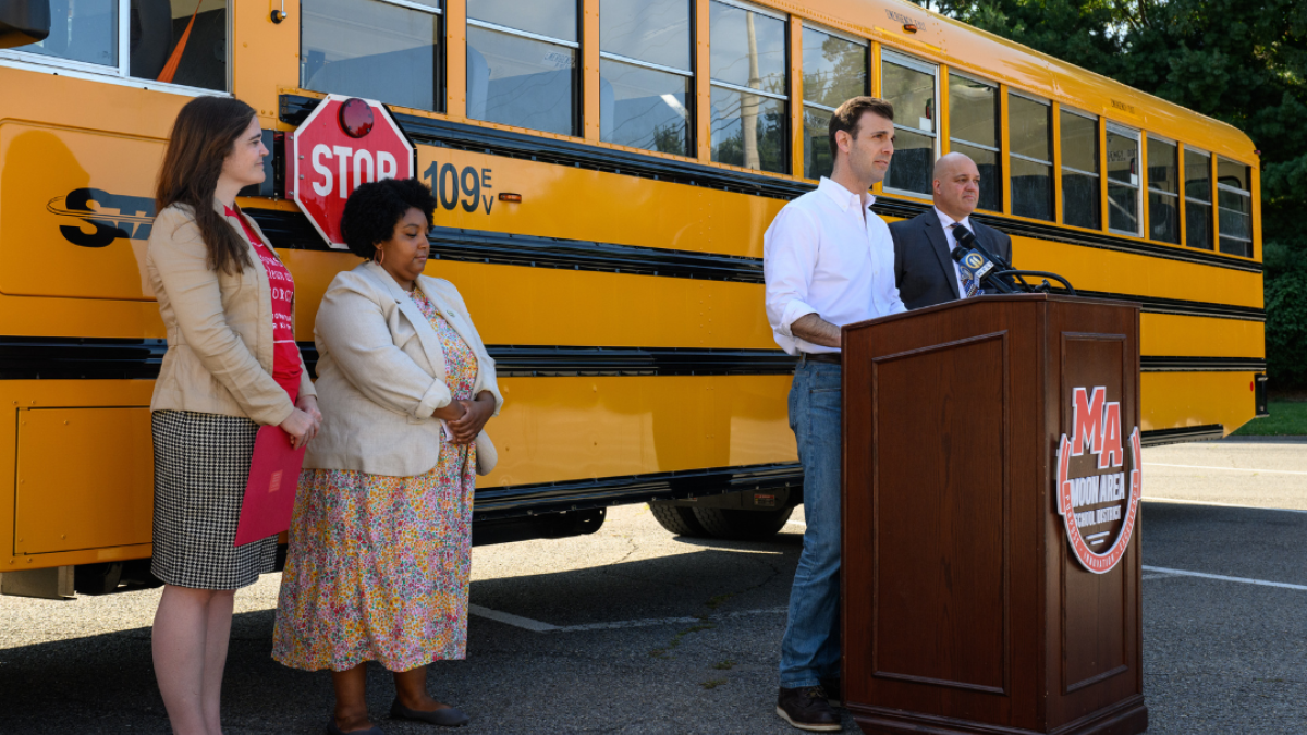 Rep. Deluzio stands behind a podium, in front of an electric school bus and gives remarks. Three other people stand behind him.