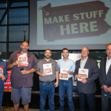 Six men stand together, holding pdfs in front of a large screen that says "MAKE STUFF HERE"
